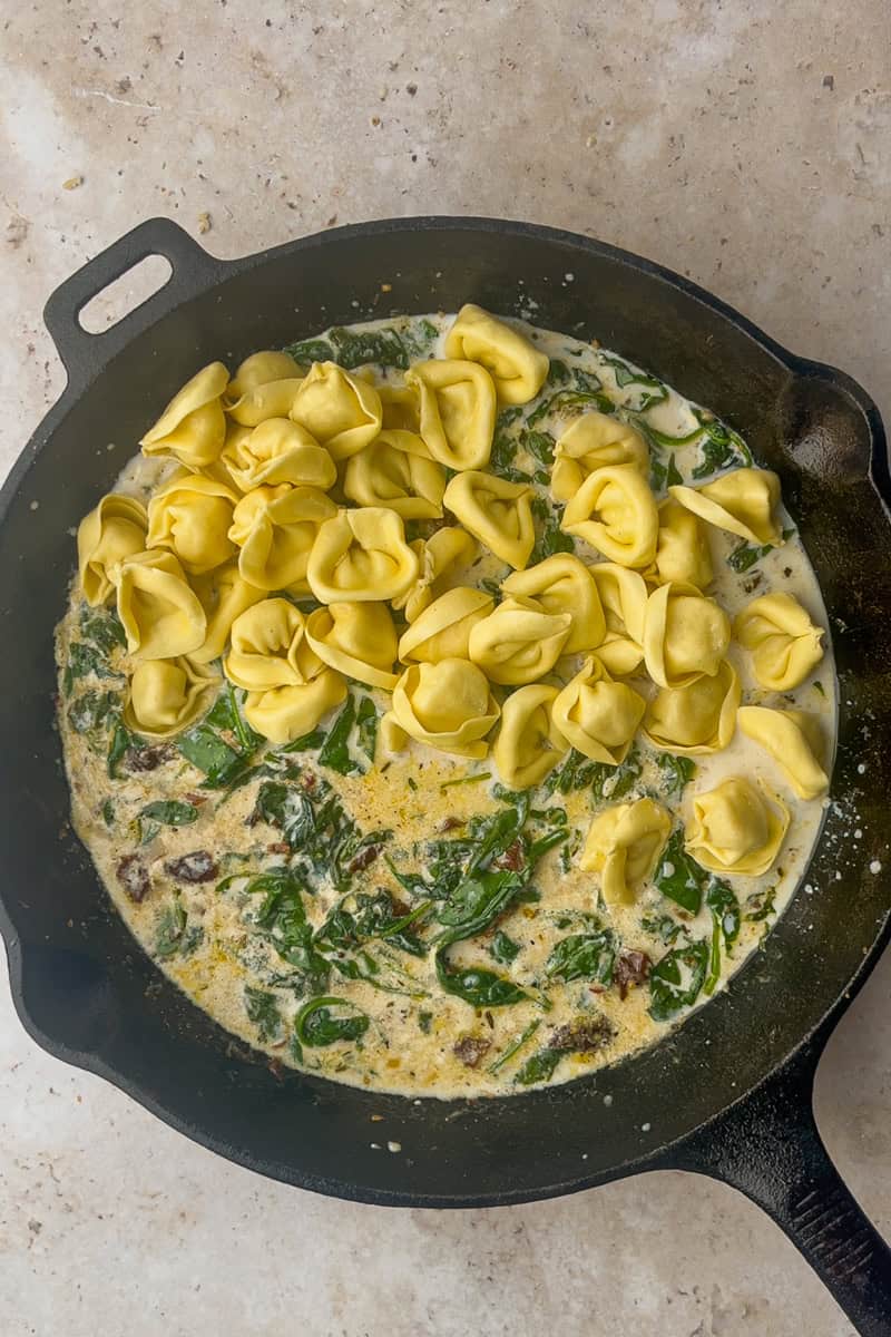 Cheese tortellini added to a creamy spinach and sun-dried tomato sauce in a cast-iron skillet.