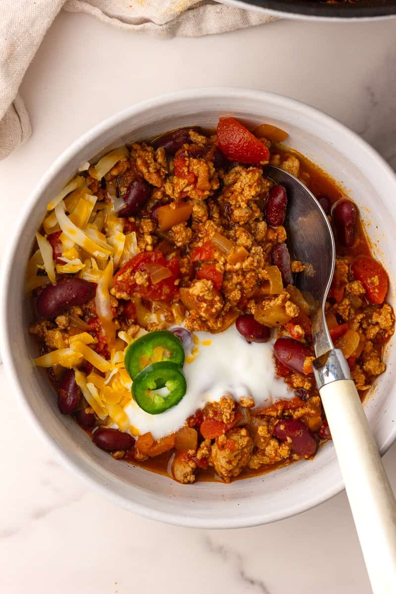 A close-up bowl of chili shows melted cheese, sour cream, jalapeño slices, and a spoon ready to serve.