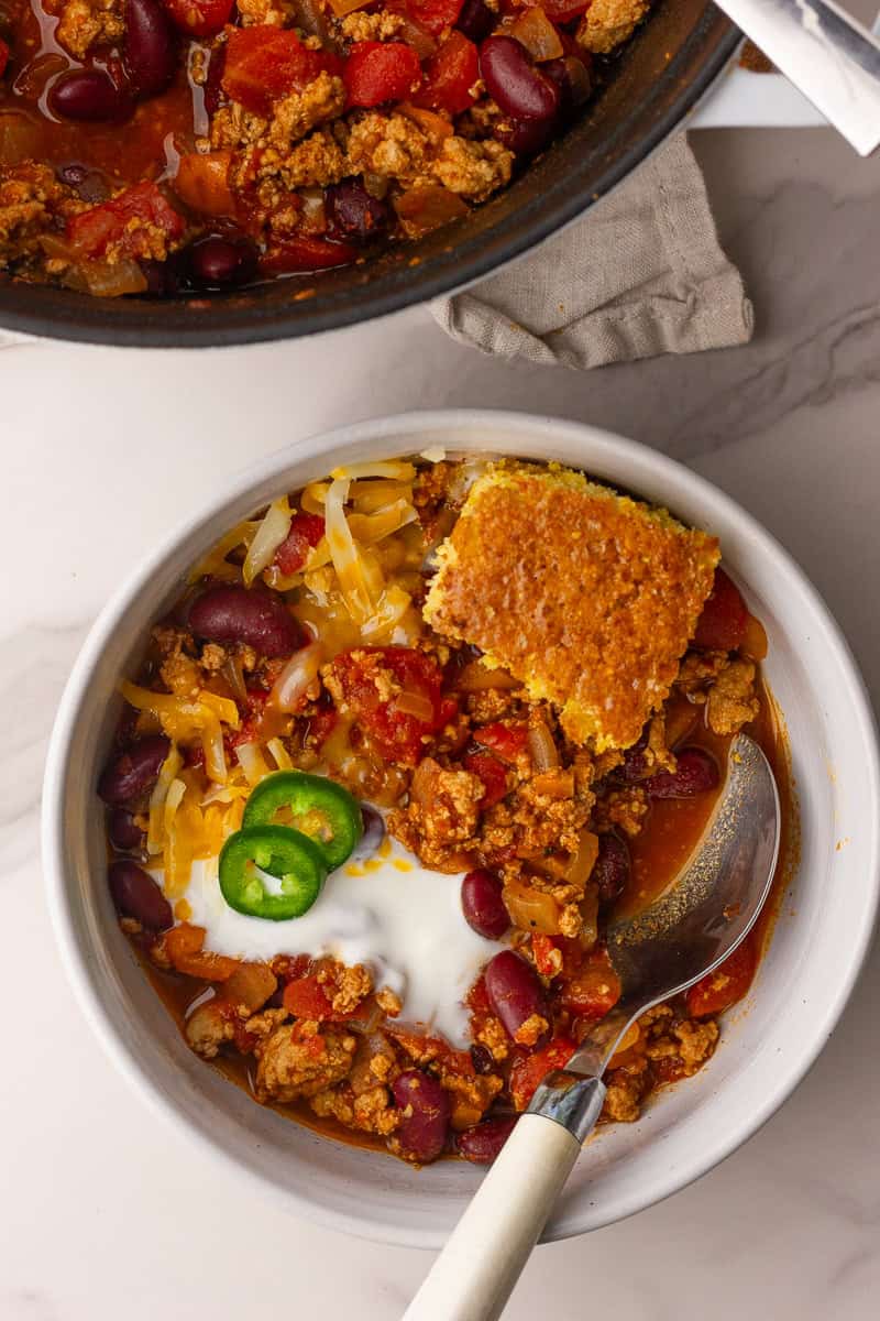 A bowl of chili is topped with shredded cheese, a dollop of yogurt or sour cream, jalapeño slices, and a wedge of cornbread.