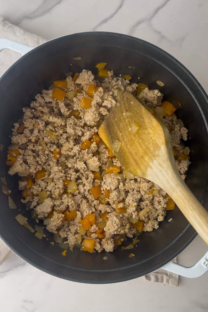 Ground meat cooks with peppers and onions in a Dutch oven as a wooden spoon stirs.