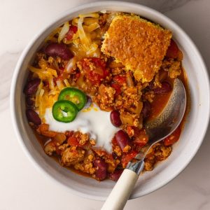 A bowl of chili is topped with shredded cheese, a dollop of yogurt or sour cream, jalapeño slices, and a wedge of cornbread.