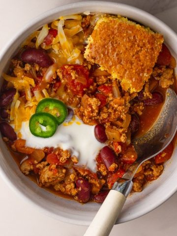 A bowl of chili is topped with shredded cheese, a dollop of yogurt or sour cream, jalapeño slices, and a wedge of cornbread.