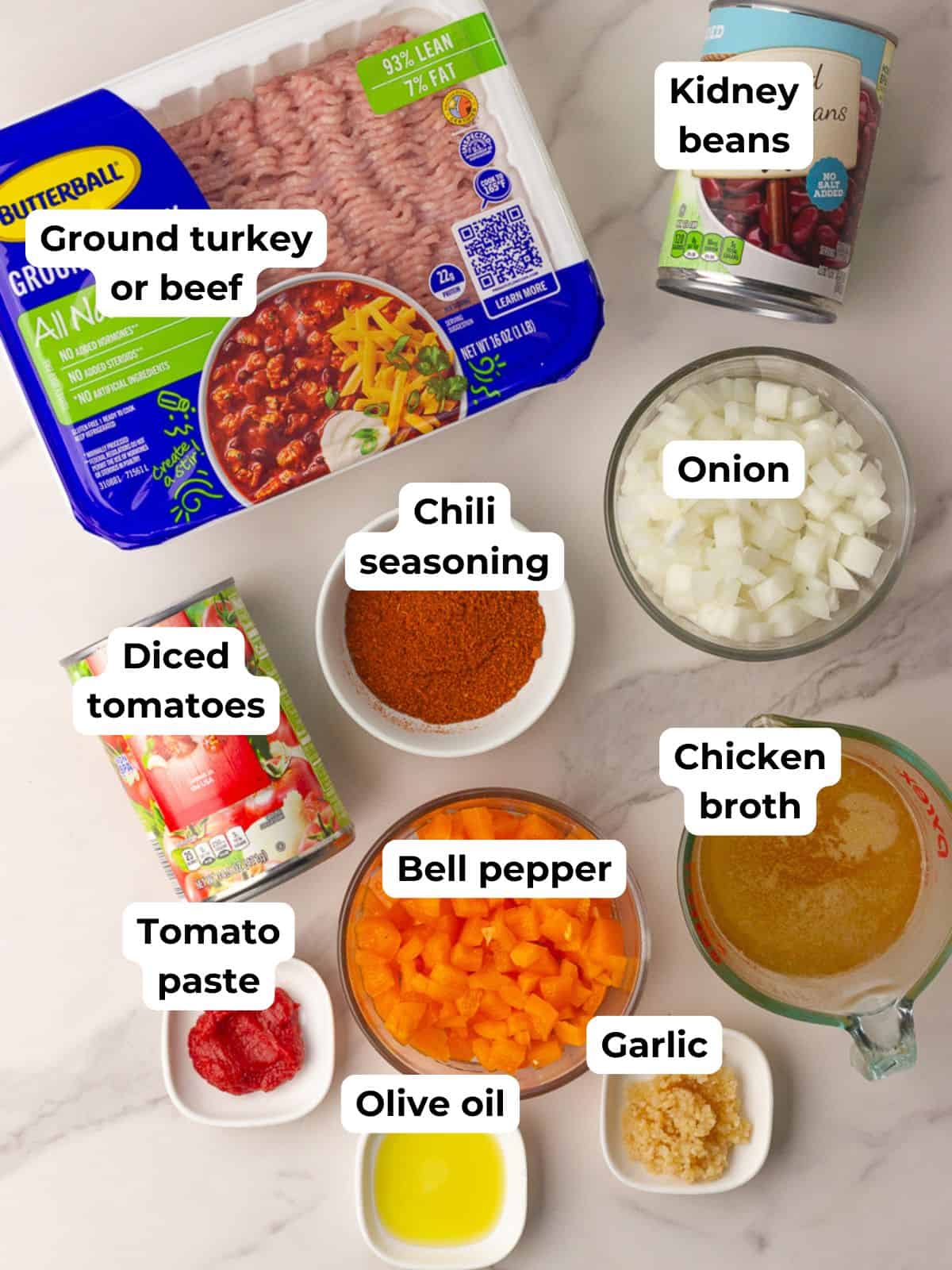 The recipe ingredients are laid out on a counter including ground turkey, kidney beans, diced tomatoes, tomato paste, bell pepper, onion, garlic, chicken broth, chili seasoning, and olive oil.