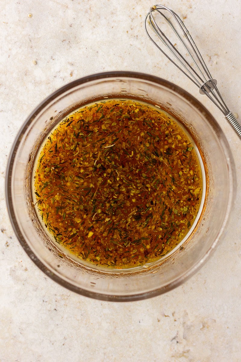 A glass bowl of maple Dijon marinade with visible herbs sits on a light countertop beside a whisk.