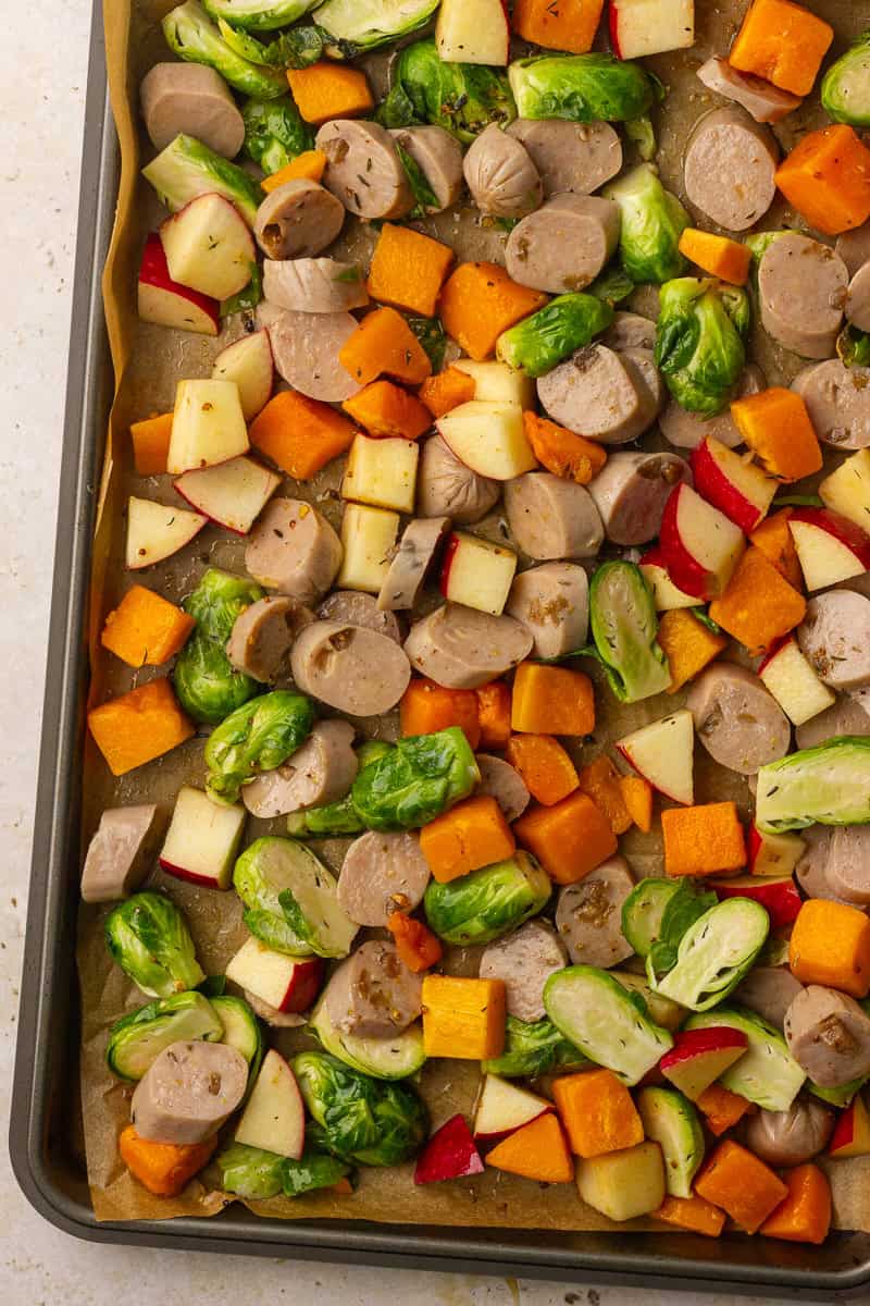 An unbaked sheet pan shows chicken sausage, butternut squash, brussels sprouts, and apple tossed in marinade and spread in a single layer.