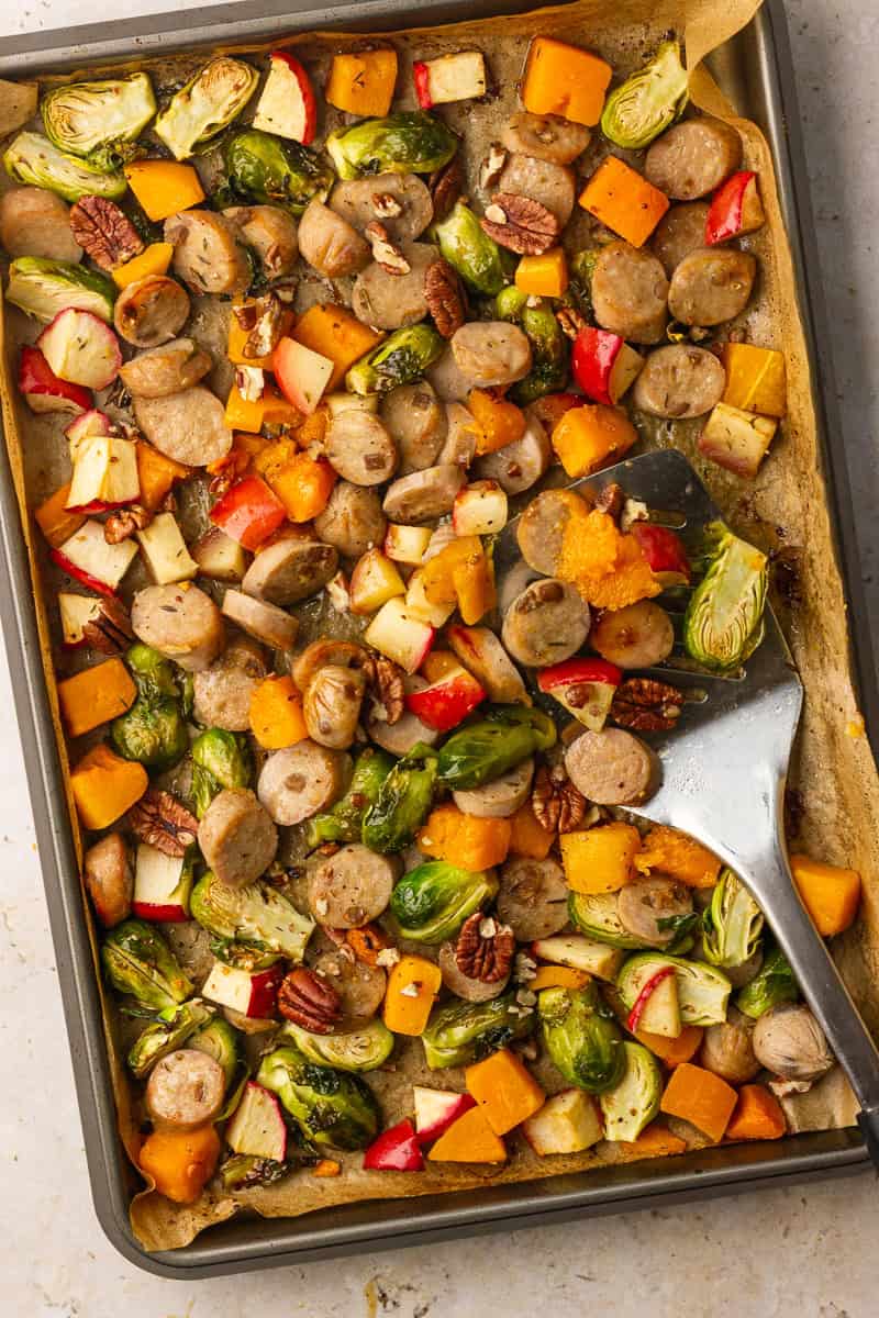 A roasted sheet pan of chicken sausage, brussels sprouts, apples, and butternut squash is sprinkled with pecans and ready to serve with a spatula.