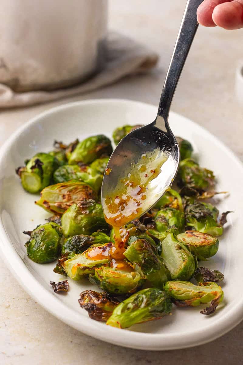 A spoon drizzles warm hot honey glaze over roasted Brussels sprouts on a white plate.