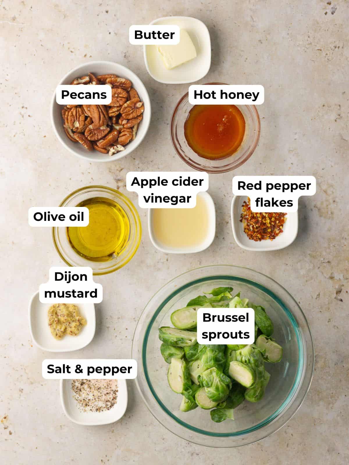 Flat lay of ingredients for hot honey Brussels sprouts including sprouts, hot honey, Dijon, apple cider vinegar, olive oil, butter, red pepper flakes, salt, pepper, and pecans.