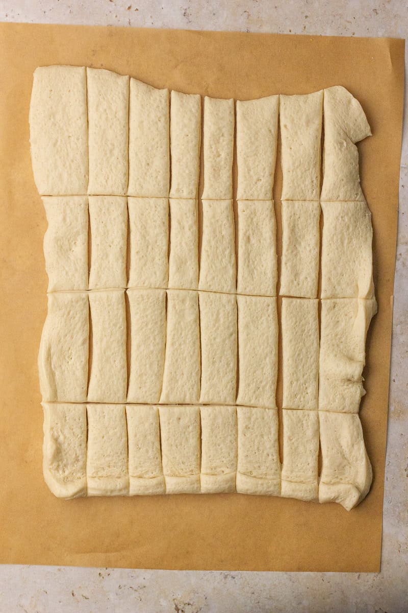 Pizza crust dough sheet cut into even strips on parchment paper, ready for wrapping.