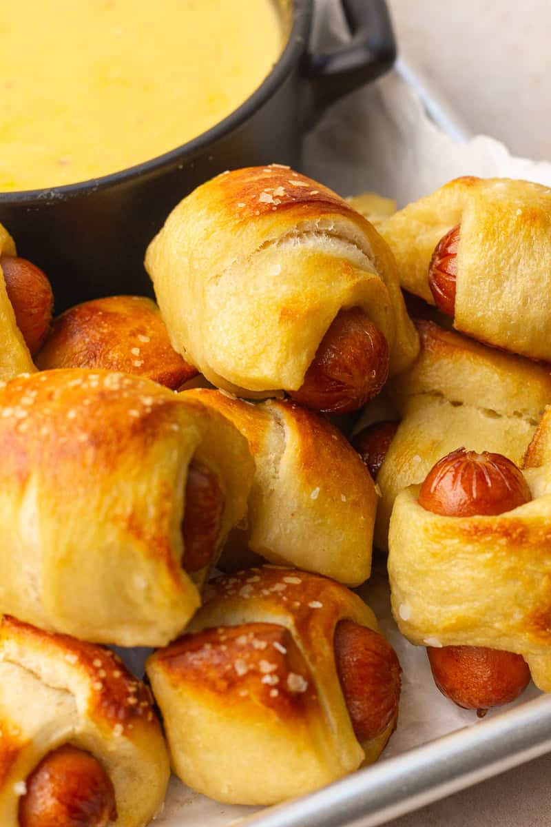 Close-up of golden mini pretzel dogs with coarse salt on top, piled next to a small bowl of creamy cheese dip.