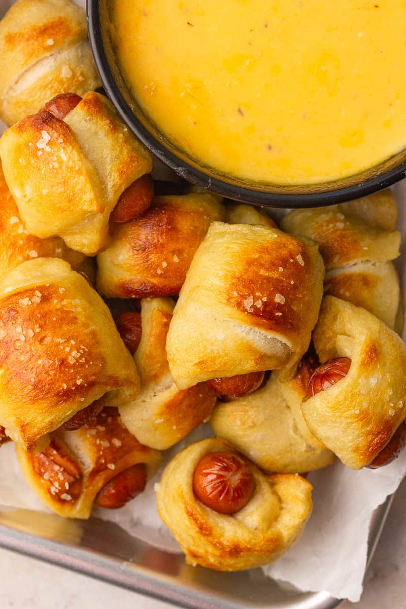 Close-up of golden-brown mini pretzel dogs sprinkled with coarse salt, served beside a bowl of cheese dip.