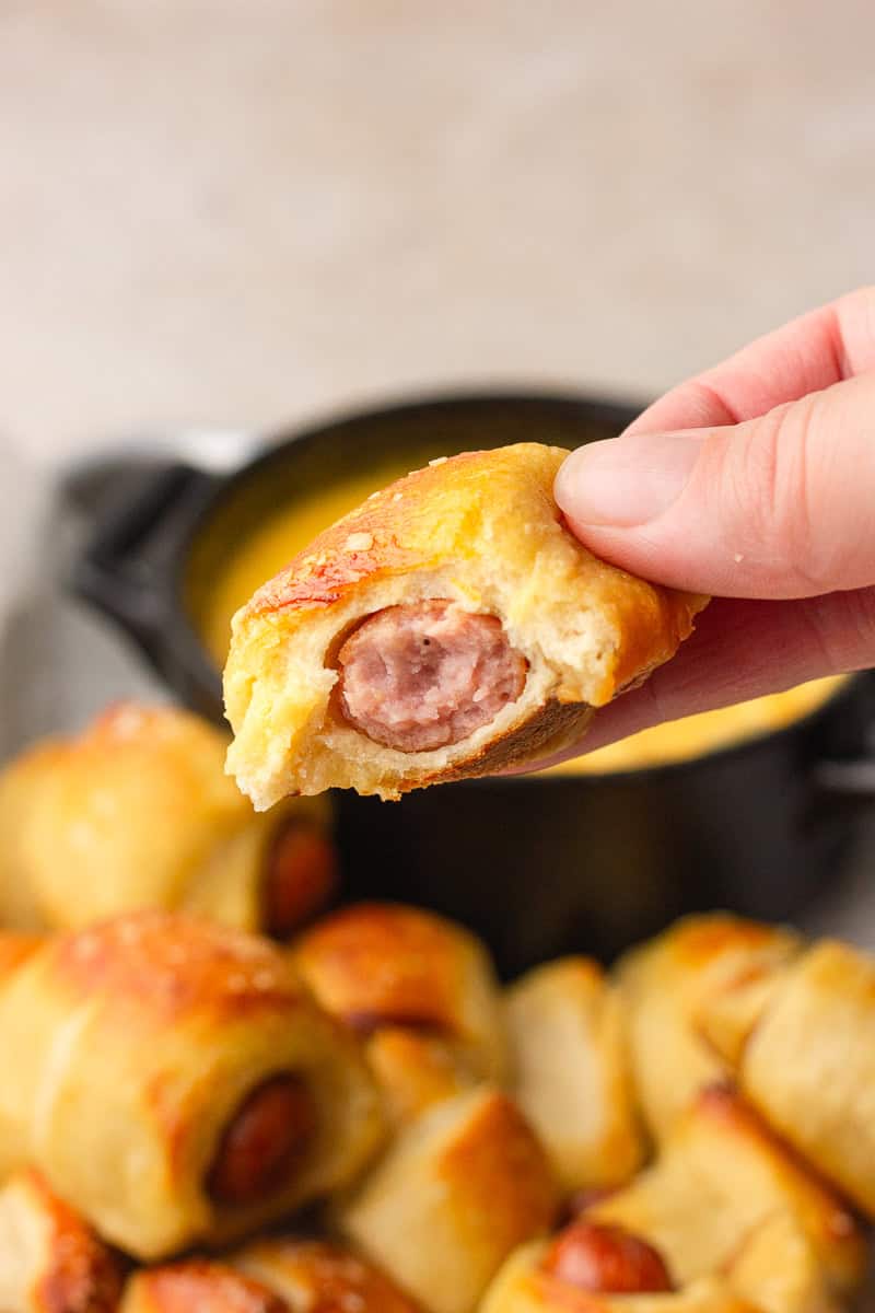 Hand holding a mini pretzel dog with a bite taken out, showing the sausage inside with cheese dip in the background.