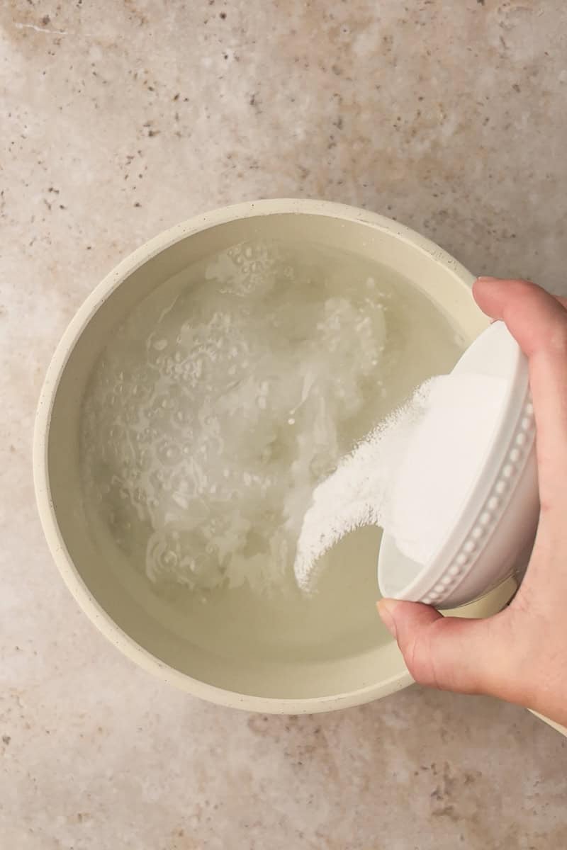 Hand pouring baking soda into a pot of boiling water to prepare the pretzel bath.