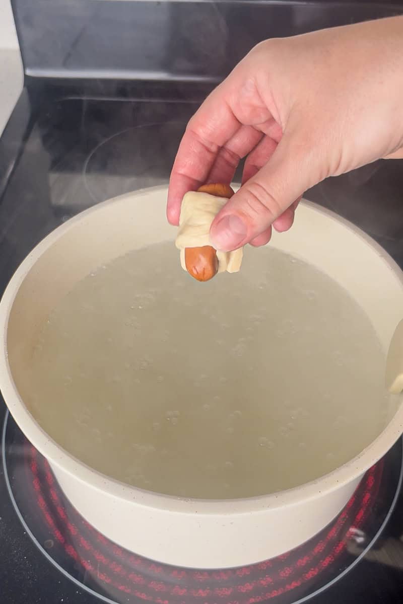 Hand holding a wrapped little smokie ready to be boiled, with dough strips in the background.
