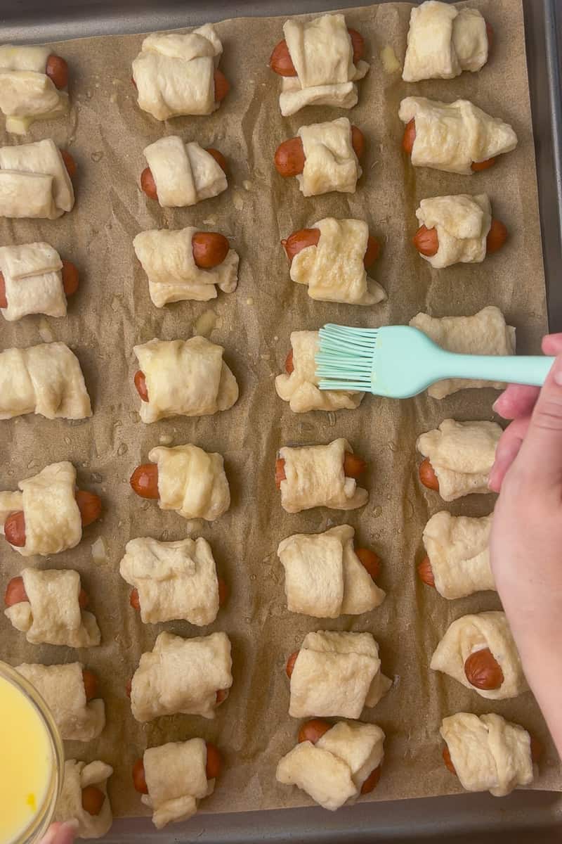Hand brushing egg wash over boiled mini pretzel dogs on a parchment-lined baking sheet.