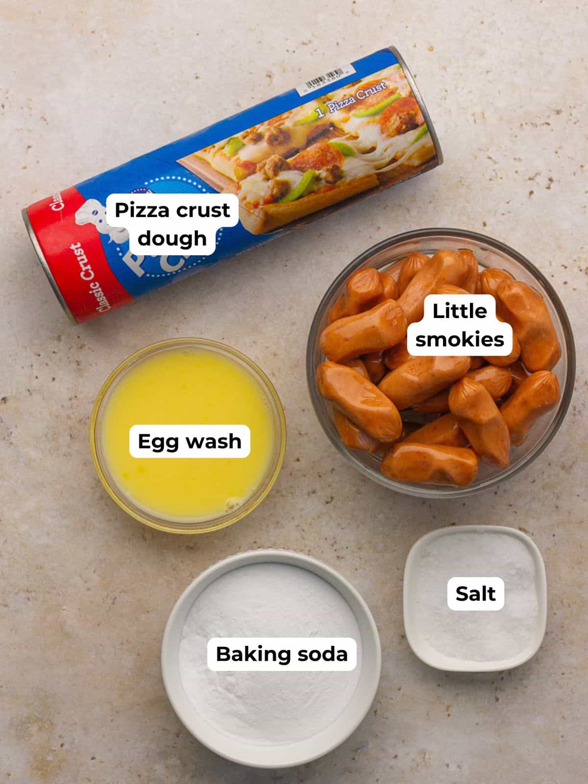 Overhead shot of pizza crust dough, little smokies, baking soda, salt, and egg wash arranged on a countertop.