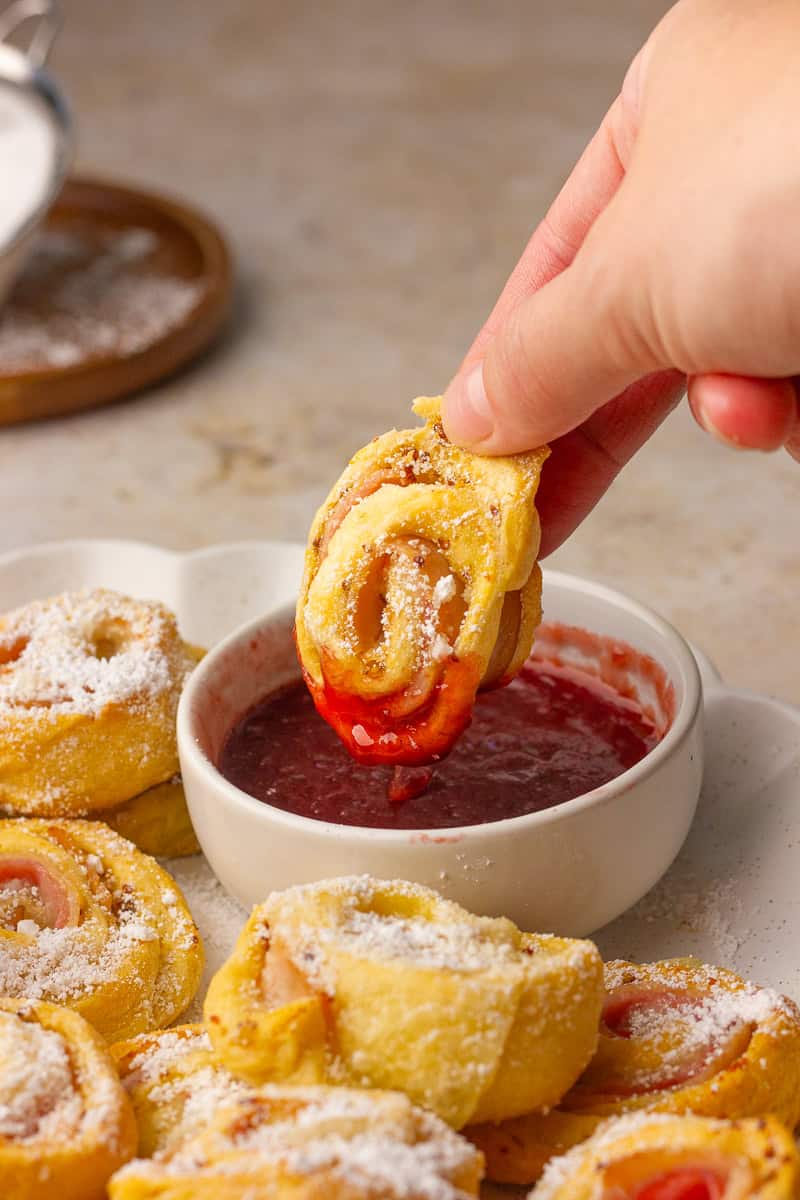 A hand dips a baked Monte Cristo pinwheel dusted with powdered sugar into a small bowl of raspberry jam.