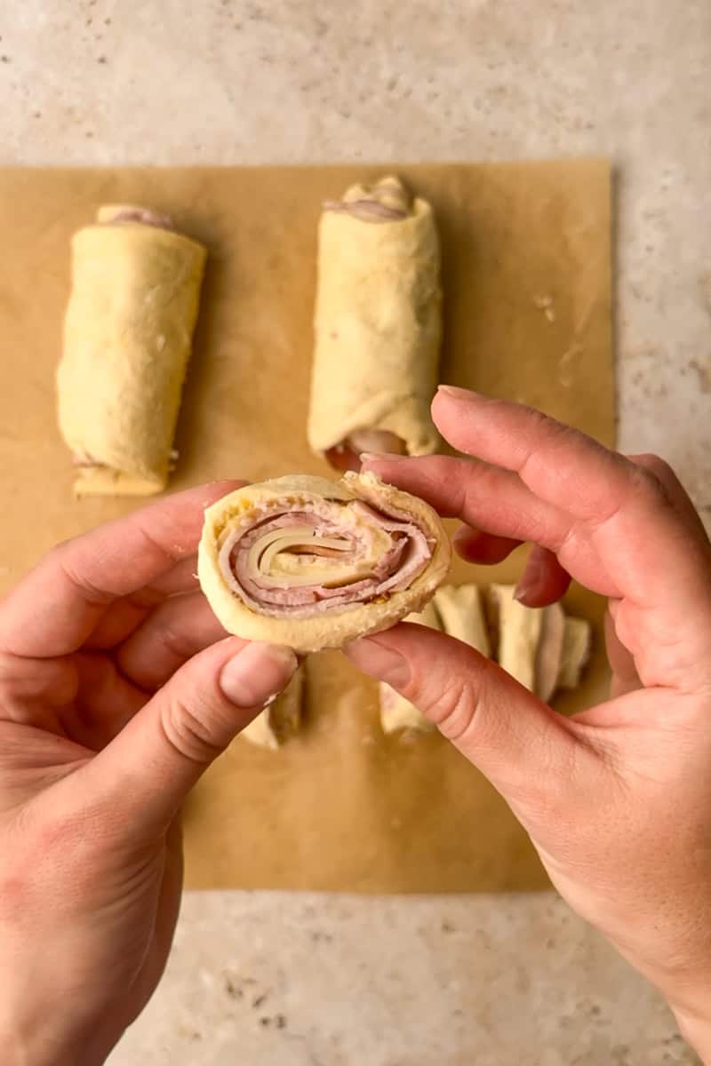 Hands hold a neatly sliced uncooked pinwheel showing thin layers of ham, turkey, and Swiss.