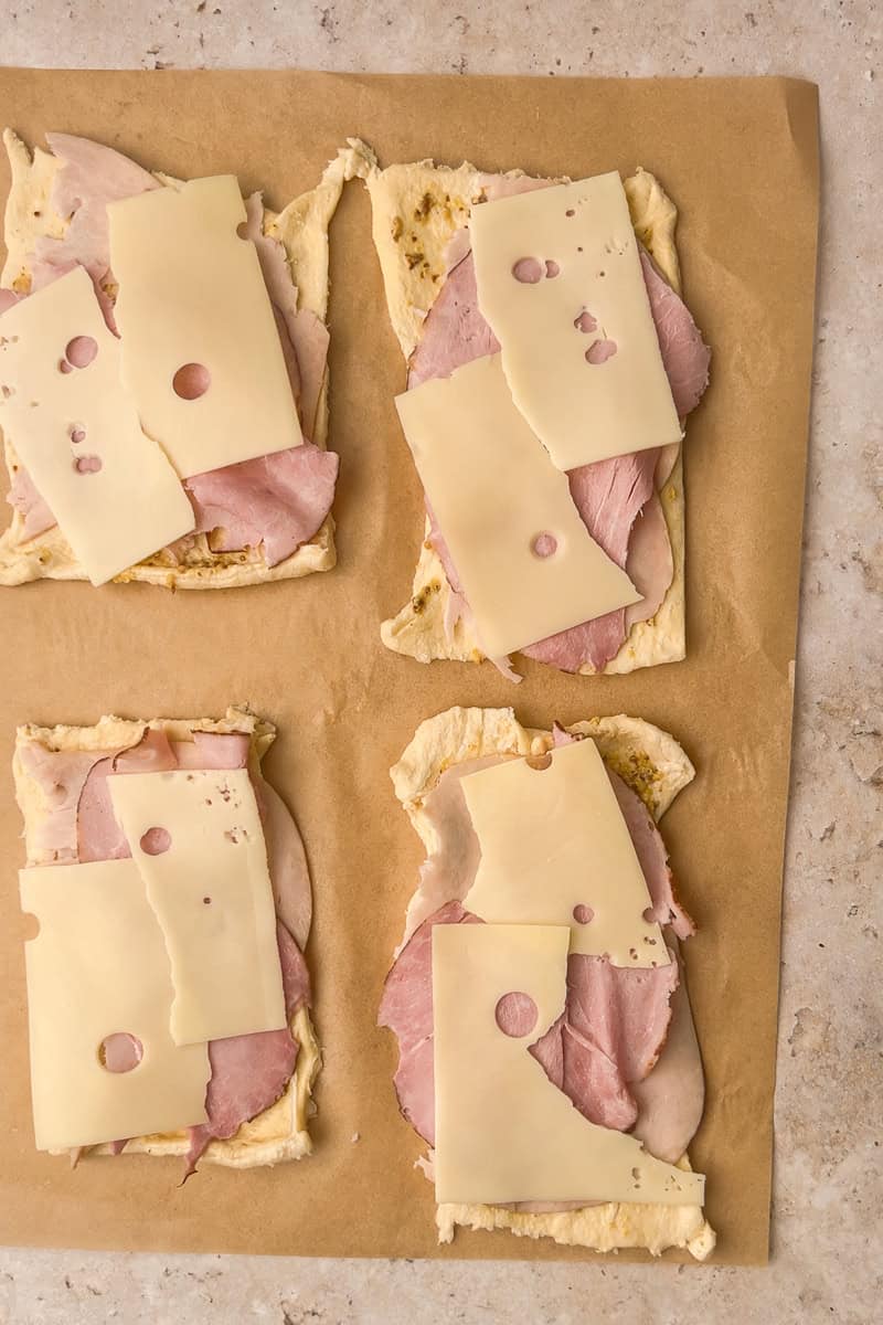 Four dough rectangles are topped with layers of ham and Swiss cheese, ready to roll.