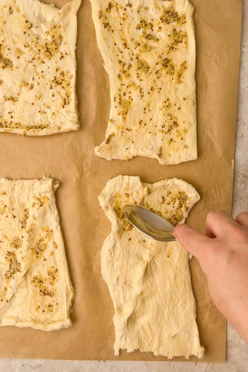 A spoon spreads a thin layer of Dijon mustard over the crescent dough rectangles.