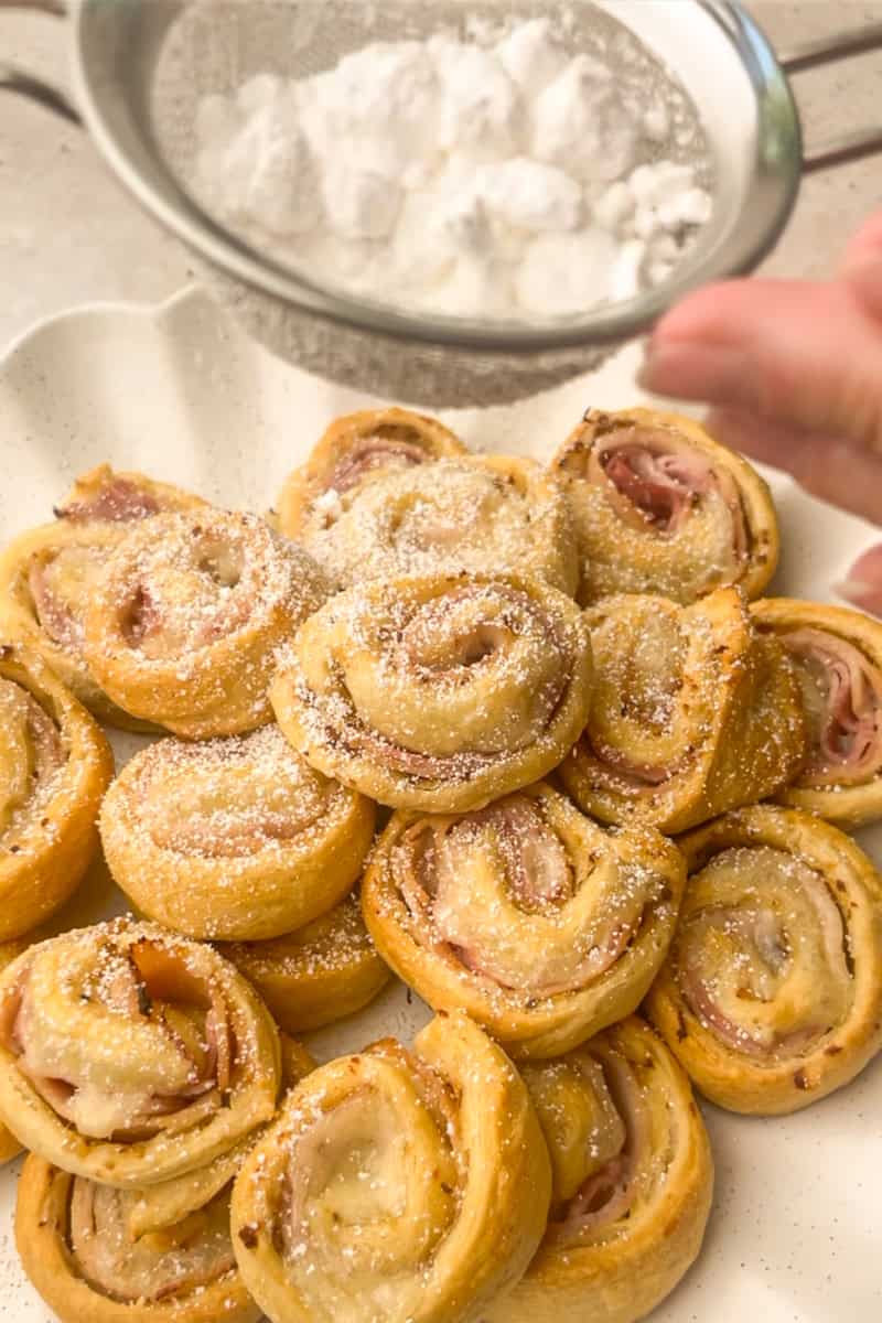 A platter of golden baked pinwheels is being dusted with powdered sugar through a sieve.