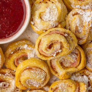 Close-up of baked Monte Cristo pinwheels sprinkled with powdered sugar beside a bowl of raspberry jam.