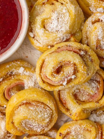 Close-up of baked Monte Cristo pinwheels sprinkled with powdered sugar beside a bowl of raspberry jam.
