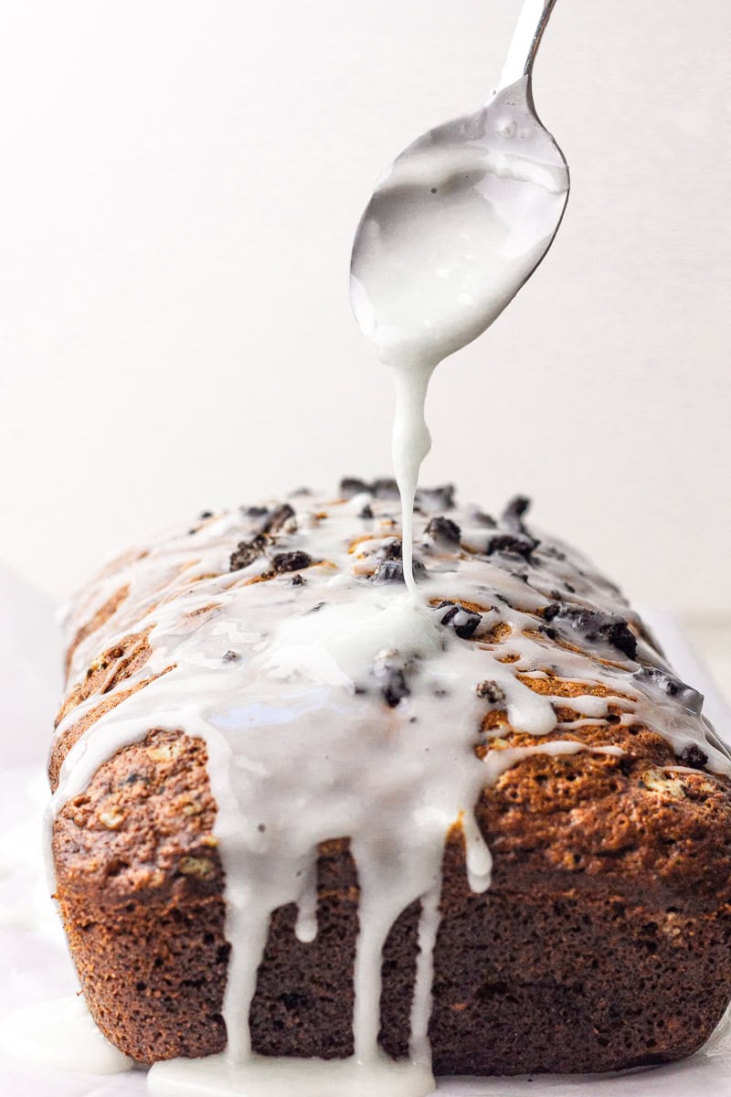 A spoon drizzles white glaze over the loaf in a close-up action shot.