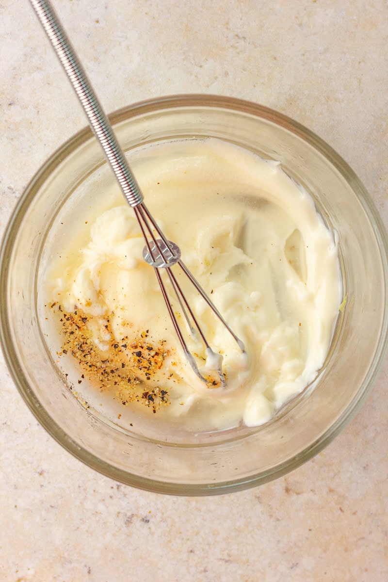 A glass bowl holds mayonnaise dressing being whisked with pepper and seasonings.