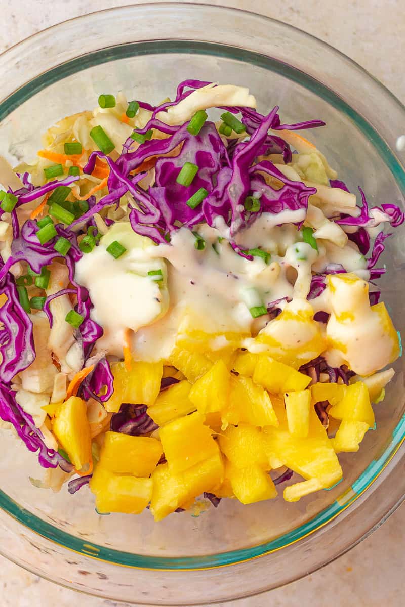 Shredded cabbage, pineapple chunks, green onions, and creamy dressing sit in a mixing bowl.
