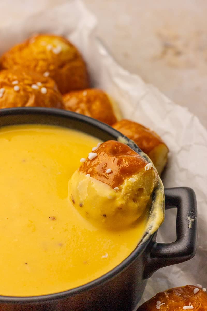 Close-up of a pretzel bite coated in melted cheese resting on the edge of a black bowl of dip.