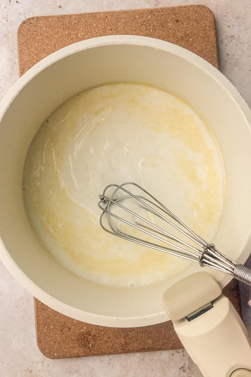 Saucepan with butter and milk mixture being whisked to start the cheese sauce base.