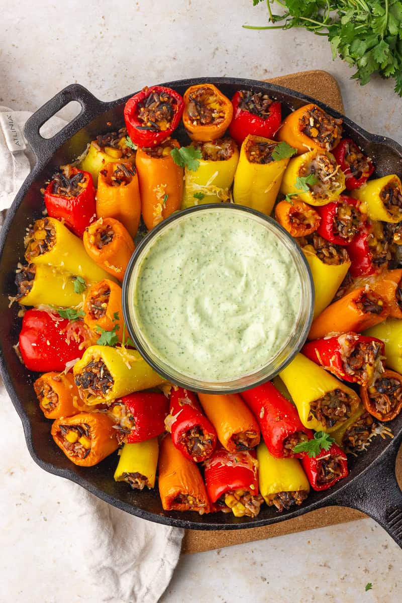 A cast-iron skillet is filled with rice-stuffed mini peppers arranged around a bowl of creamy green dip.