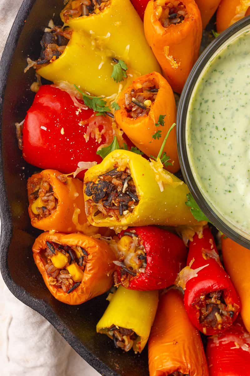A close-up shows baked mini peppers stuffed with rice and corn beside the dip bowl in a cast-iron skillet.