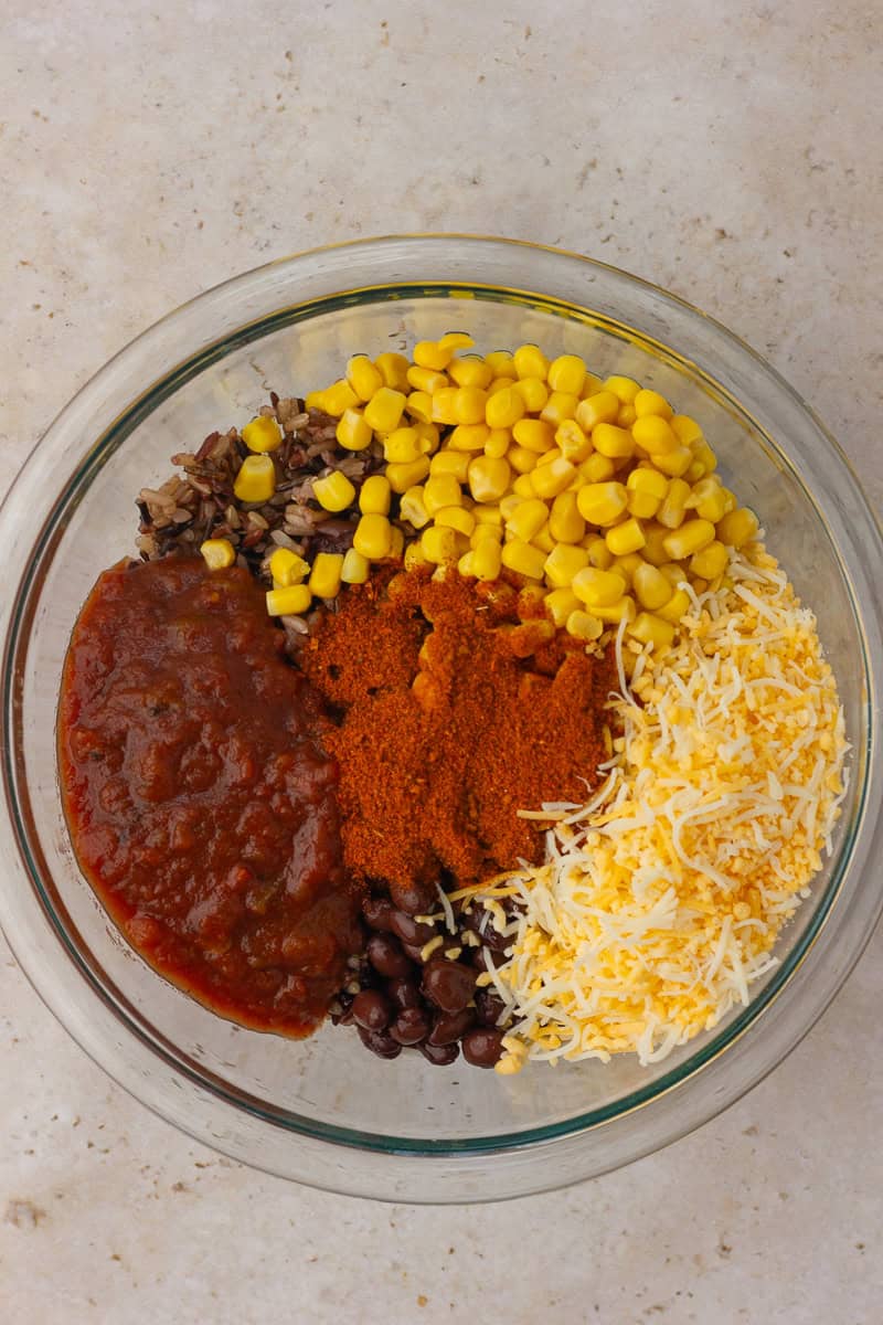 A mixing bowl contains wild rice, black beans, corn, salsa, taco seasoning, and shredded cheese before stirring.