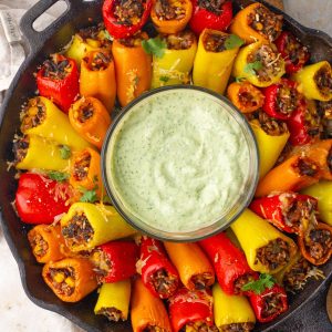 A cast-iron skillet holds colorful rice-stuffed mini peppers arranged around a bowl of creamy green dip.