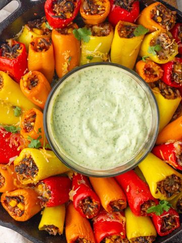 A cast-iron skillet holds colorful rice-stuffed mini peppers arranged around a bowl of creamy green dip.