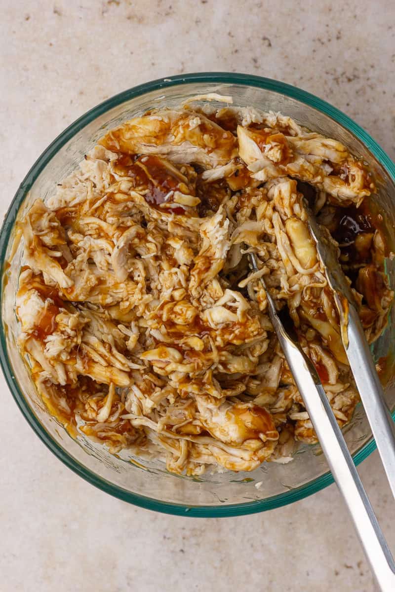 A glass bowl of shredded chicken coated in teriyaki sauce with metal tongs.
