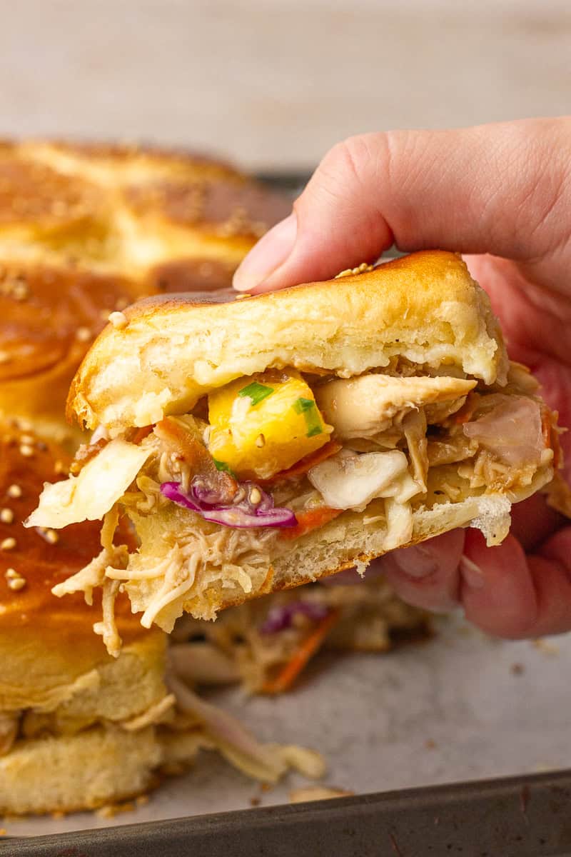A hand holds a teriyaki chicken slider on a Hawaiian roll with pineapple slaw and melted cheese.