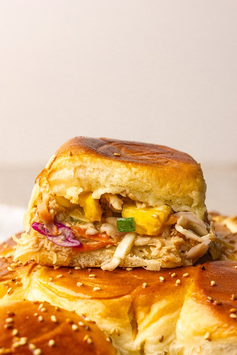A close-up shows a single teriyaki chicken slider with pineapple and slaw perched on the sesame-topped rolls.
