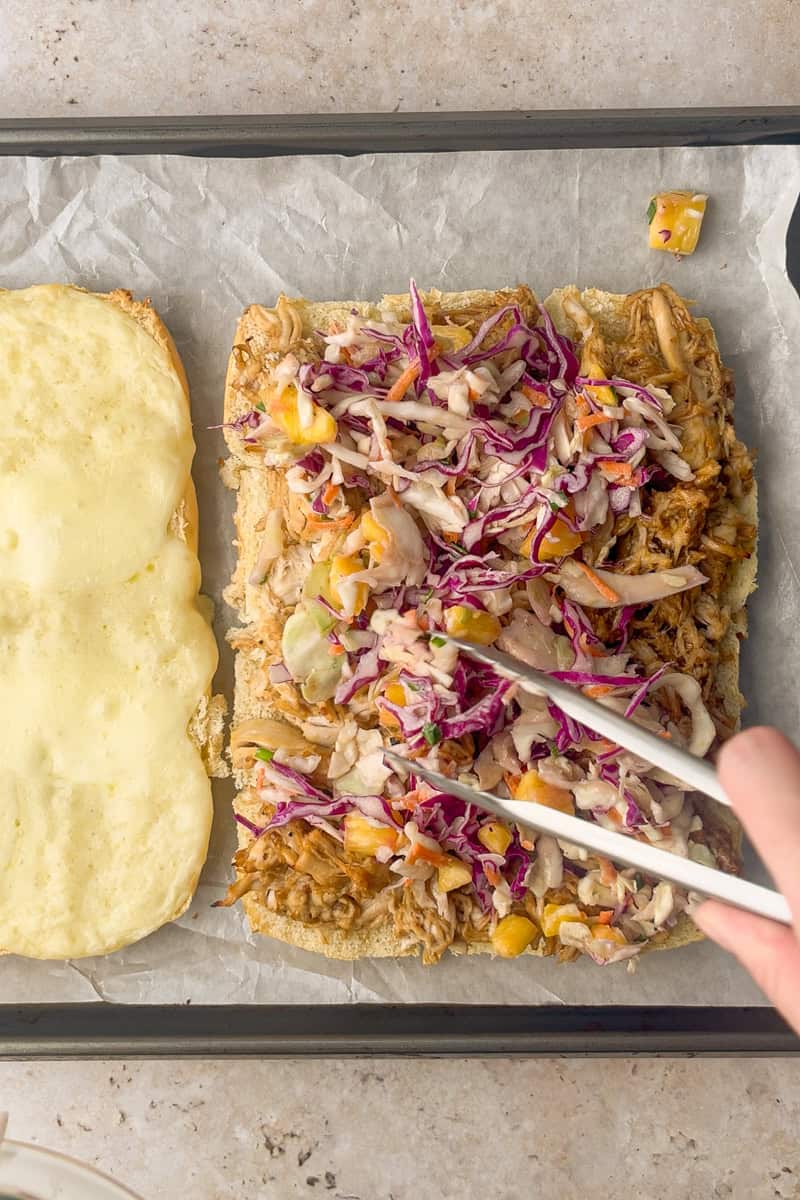 Tongs add pineapple coleslaw over teriyaki chicken on the bottom roll slab beside the melted-cheese top slab.