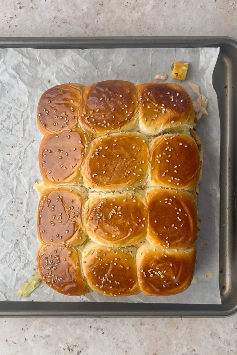 A full slab of Hawaiian rolls sits on a sheet pan brushed with butter and sprinkled with sesame seeds.
