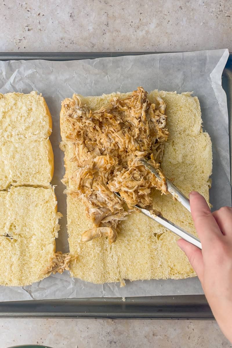Shredded teriyaki chicken being placed on the bottom layer of rolls with tongs.