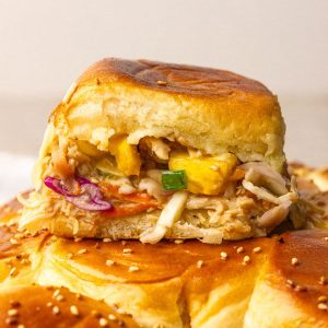 A close-up slider with pineapple and slaw rests on the sesame-topped roll slab.