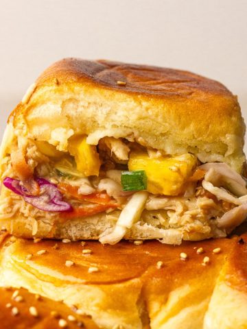 A close-up slider with pineapple and slaw rests on the sesame-topped roll slab.