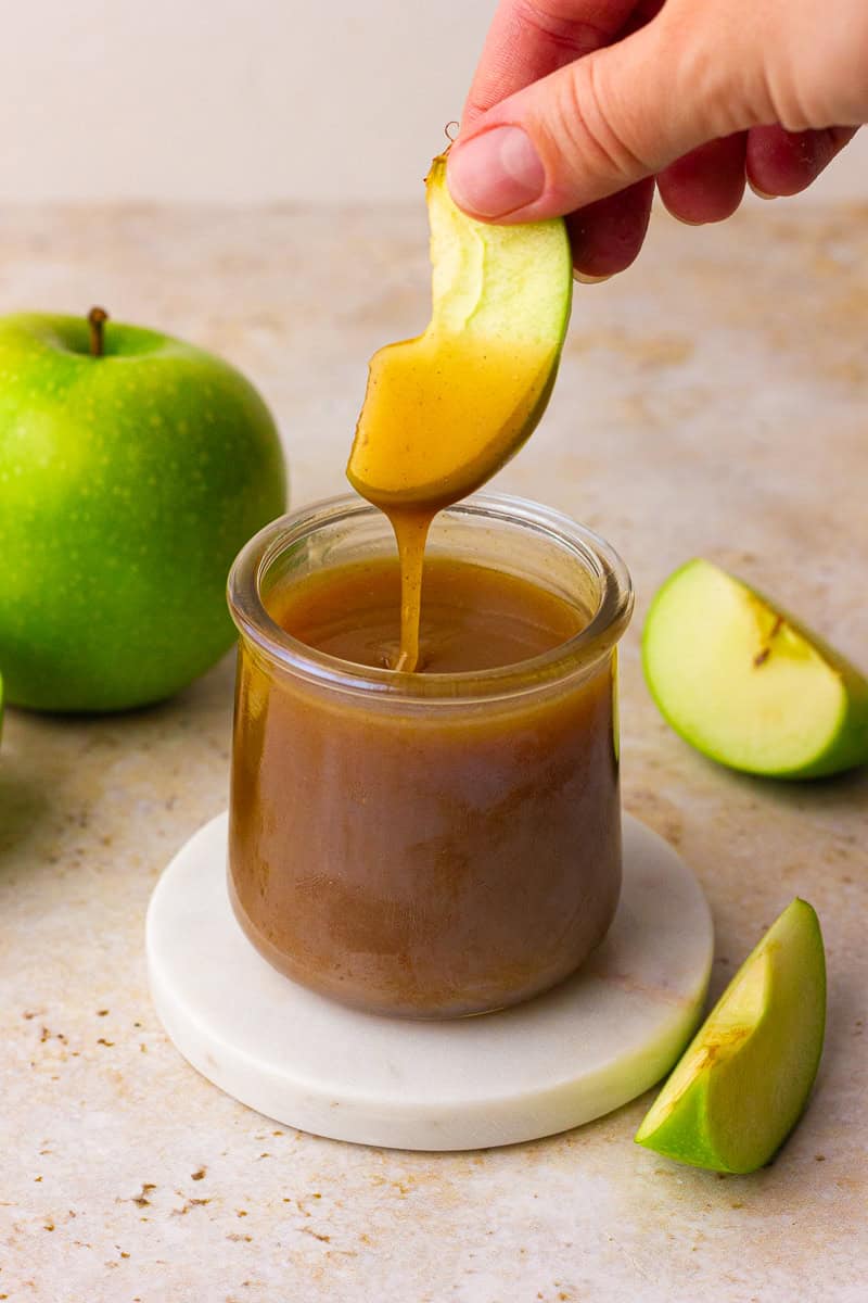 A hand dips a green apple slice into a small jar of apple cider caramel sauce as the caramel drips back into the jar.