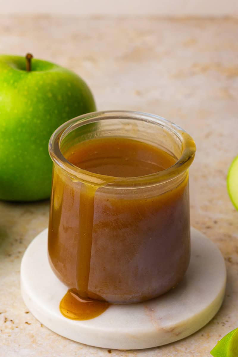 A glass jar of apple cider caramel sauce sits on a marble coaster with a caramel drip down the side and a green apple behind it.