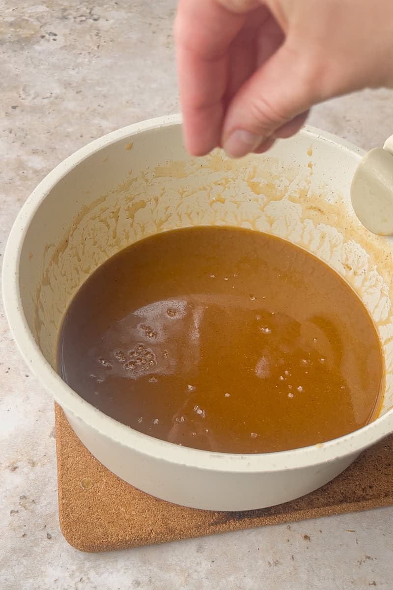 A hand sprinkles flaky salt into a pot of finished apple cider caramel sauce.