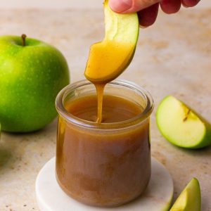 A green apple slice is dipped into a jar of apple cider caramel sauce with a visible drip.