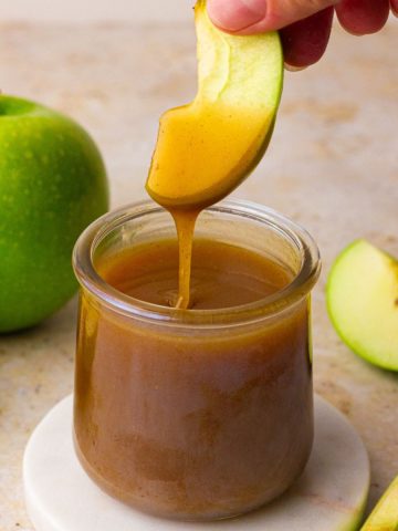 A green apple slice is dipped into a jar of apple cider caramel sauce with a visible drip.
